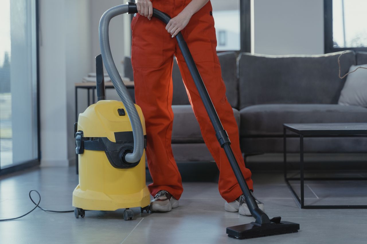 Un nettoyeur professionnel en combinaison rouge aspire un salon contemporain avec un aspirateur jaune.