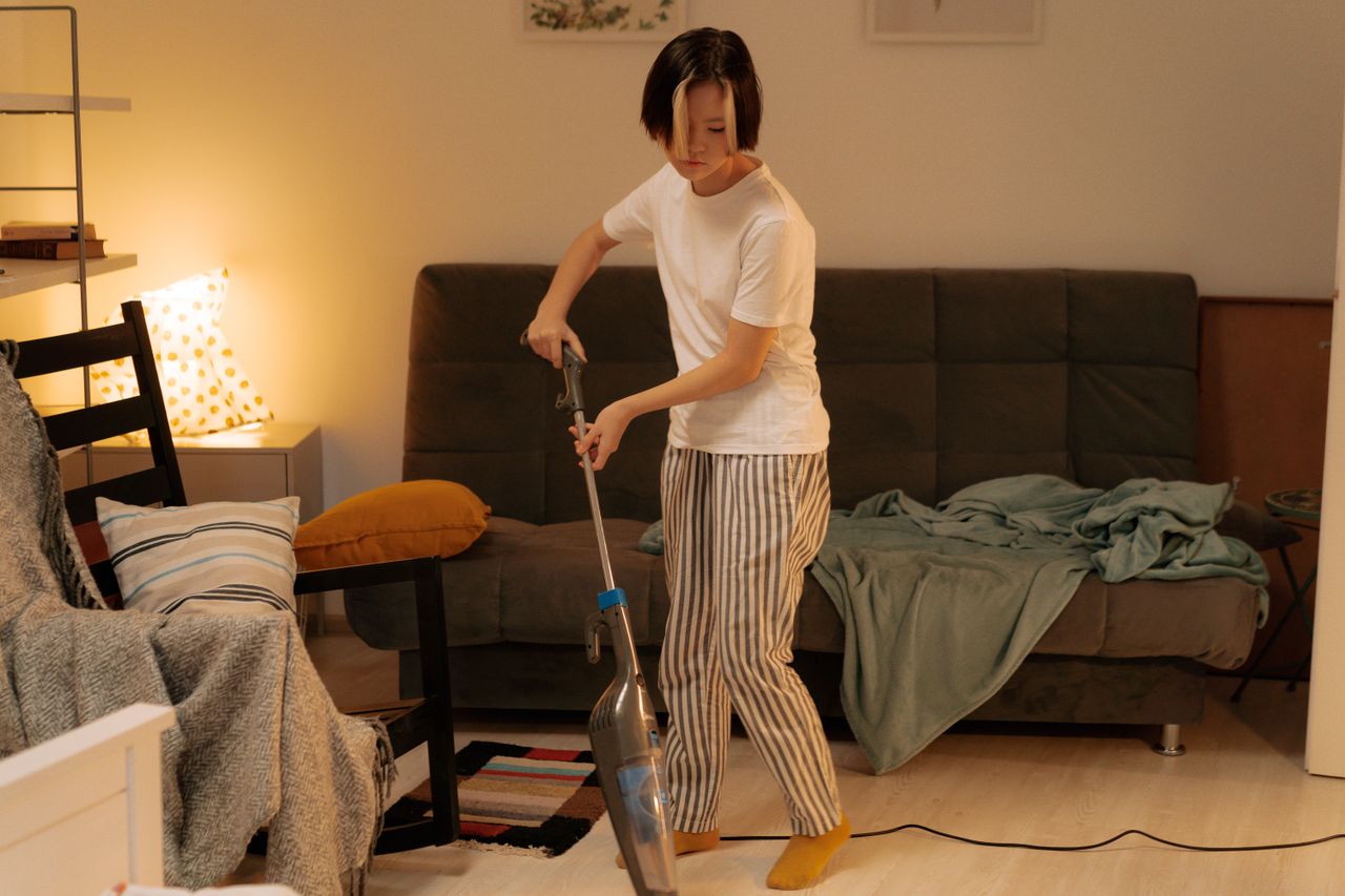 An Asian woman is cleaning a cluttered living room at home with a vacuum cleaner, making the space neat and orderly.